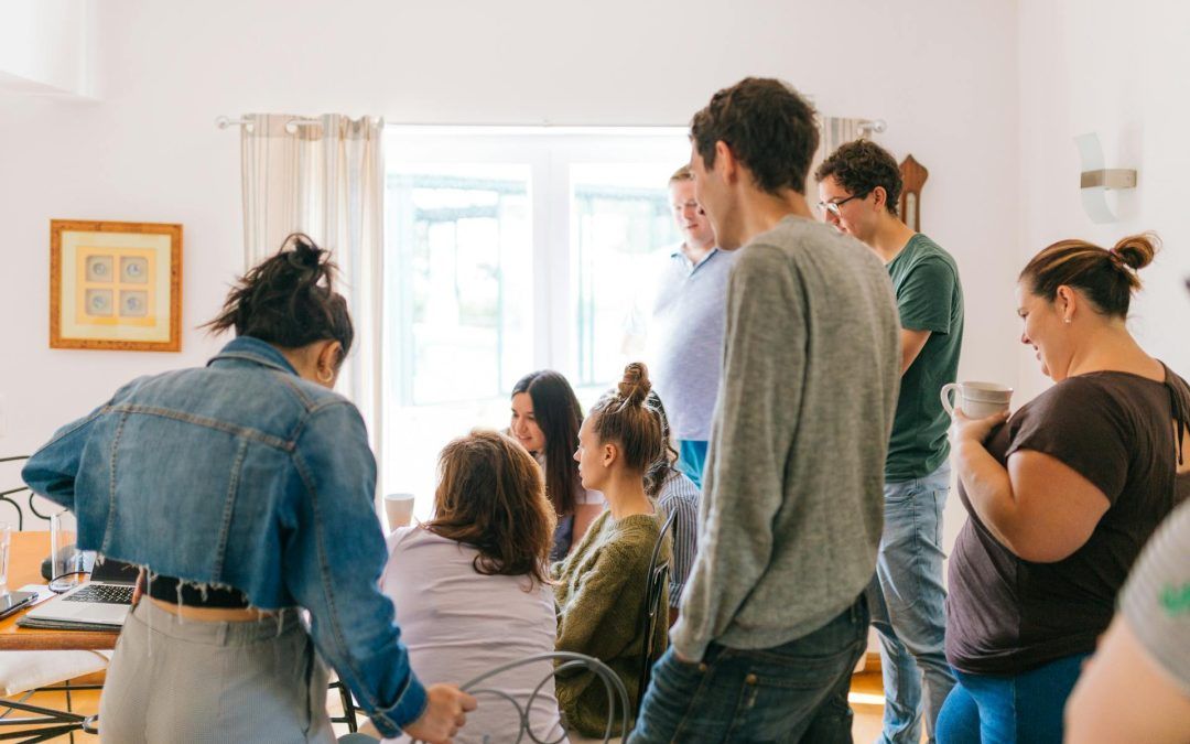 Gruppe von Erwachsenen steht und sitzt in einem hellen Raum und führt eine Diskussion oder ein Seminar durch. Eine Person sitzt mit dem Rücken zur Kamera und zeigt auf etwas, während andere aufmerksam zuhören.