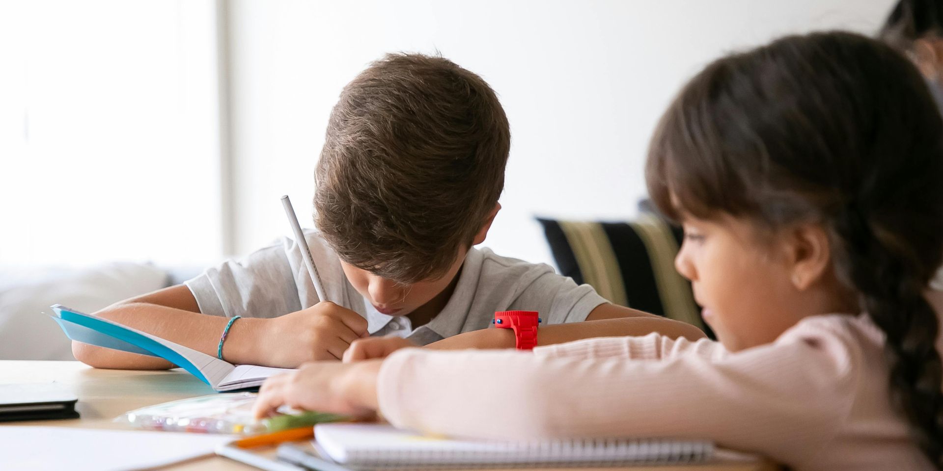 Zwei Kinder sitzen konzentriert an einem Tisch und arbeiten in Heften. Ein Kind hält einen Stift in der Hand, das andere blättert in einem Heft. Auf dem Tisch liegen Schreibmaterialien wie Stifte und ein Lineal.