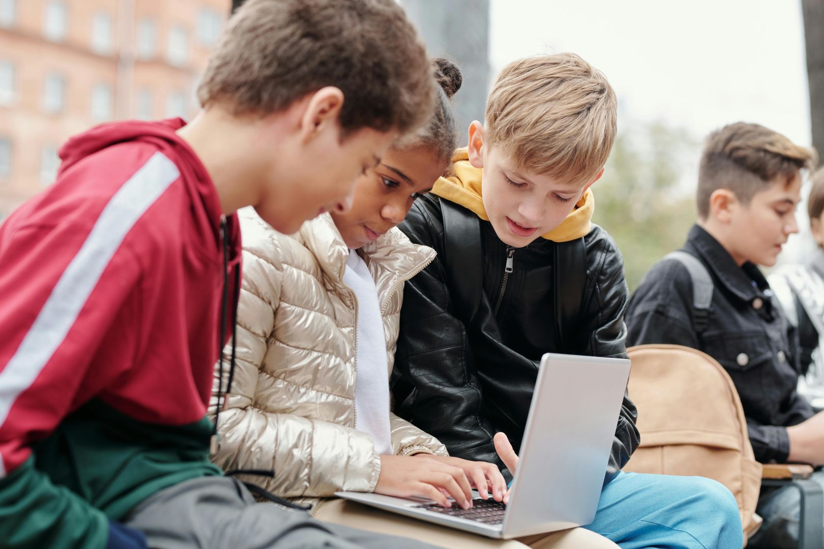 Drei Jugendliche sitzen nebeneinander auf einer Bank und schauen konzentriert auf den Bildschirm eines Laptops. Im Hintergrund sind weitere Jugendliche zu sehen.