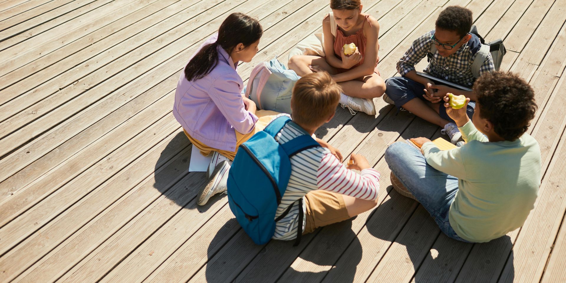 Fünf Kinder sitzen im Kreis auf einem Holzdeck und essen gemeinsam. Zwei Kinder essen Apfel, ein anderes hat einen blauen Rucksack auf dem Rücken, eines ein Tablet in den Händen. Die Szene zeigt eine entspannte, soziale Situation unter Kindern.