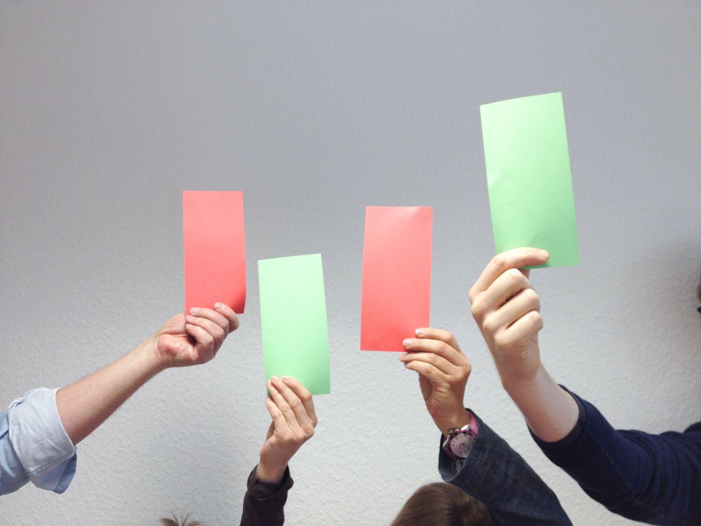Vier Hände halten abwechselnd rote und grüne Karten hoch, die an eine Ampelregelung erinnern. Zwei rote und zwei grüne Karten sind zu sehen.