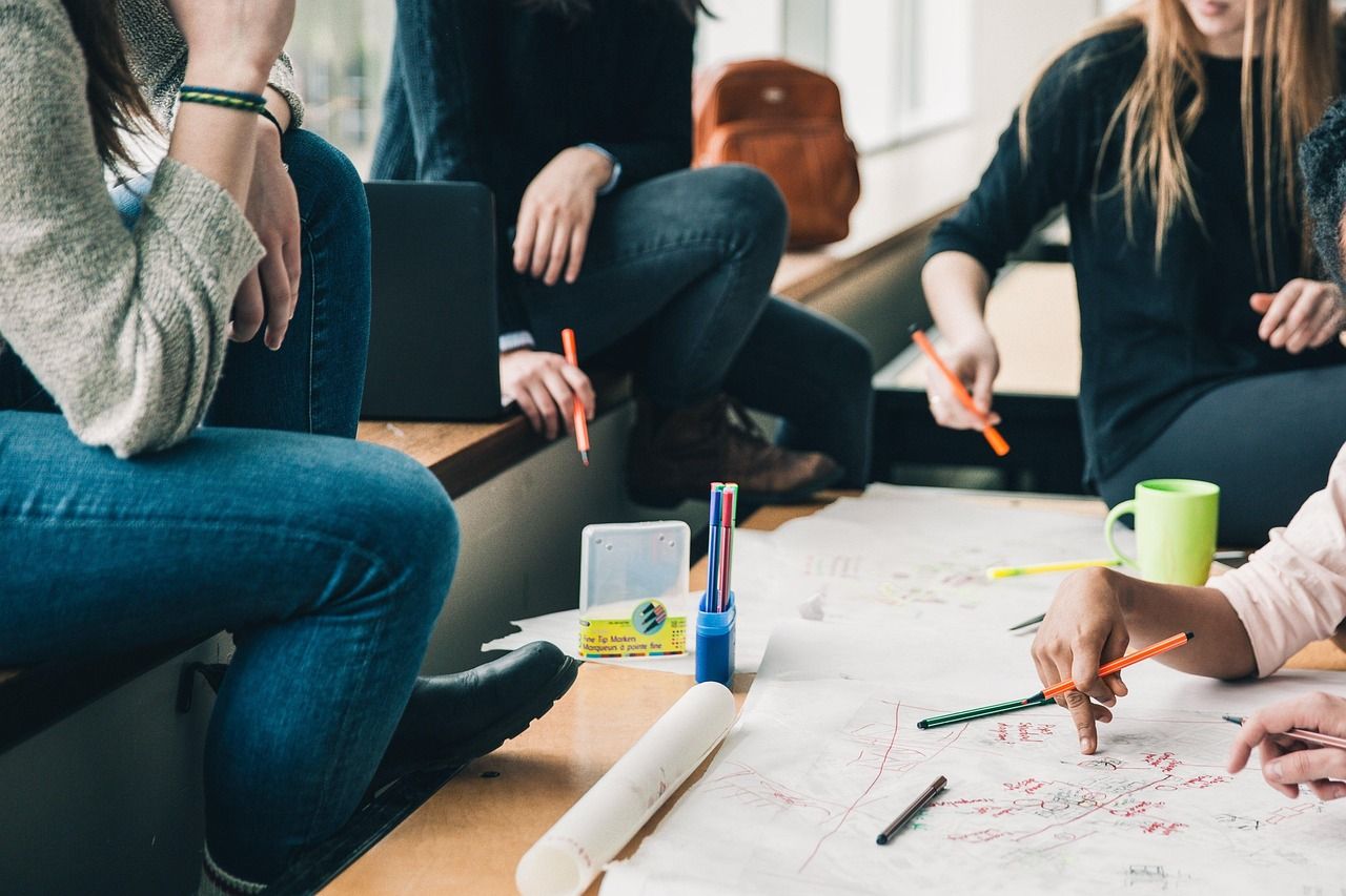 Gruppe von Personen sitzt im Kreis an einem Tisch und arbeitet gemeinsam an einem großen Blatt Papier. Auf dem Tisch liegen Stifte, Textmarker und eine Tasse. Die Szene zeigt eine kreative Gruppenarbeit oder ein Brainstorming.