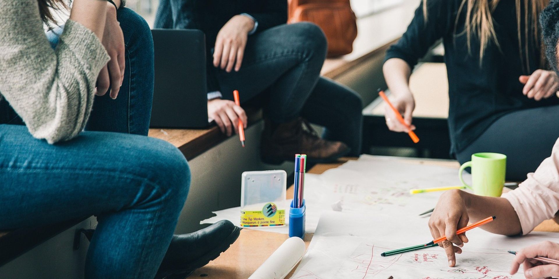 Gruppe von Personen sitzt im Kreis an einem Tisch und arbeitet gemeinsam an einem großen Blatt Papier. Auf dem Tisch liegen Stifte, Textmarker und eine Tasse. Die Szene zeigt eine kreative Gruppenarbeit oder ein Brainstorming.