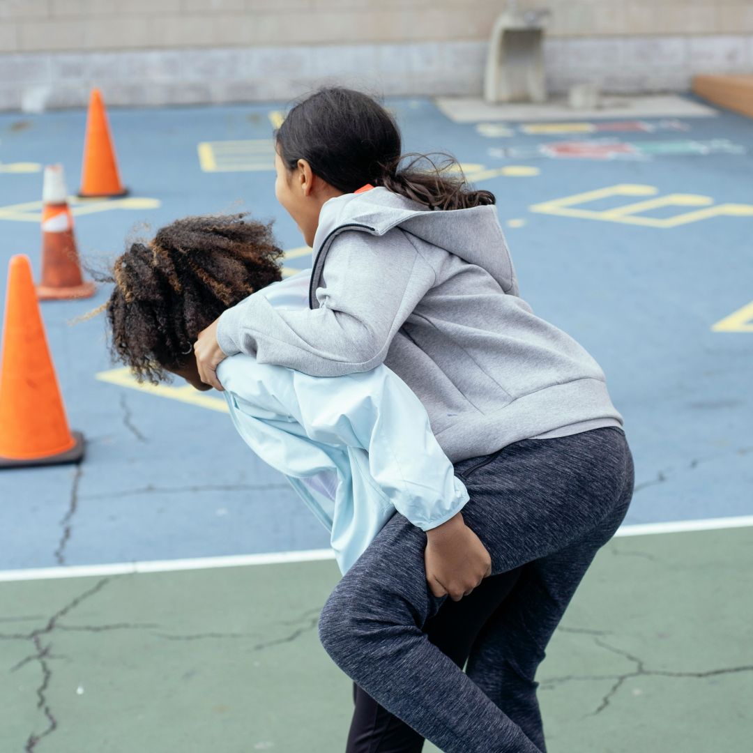 Zwei Kinder auf einem Schulhof, eines trägt das andere huckepack. Beide lachen und wirken fröhlich. Im Hintergrund sind orange Verkehrshütchen und ein Schulgebäude zu sehen.